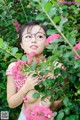 A woman wearing glasses standing in front of a bush of flowers.